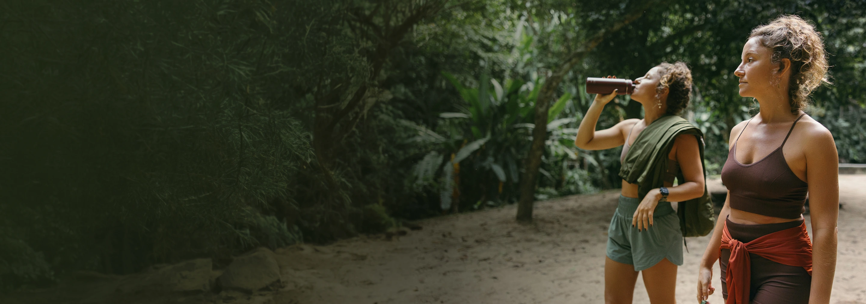 Two women in activewear outdoors, one drinking from a bottle, surrounded by green trees.