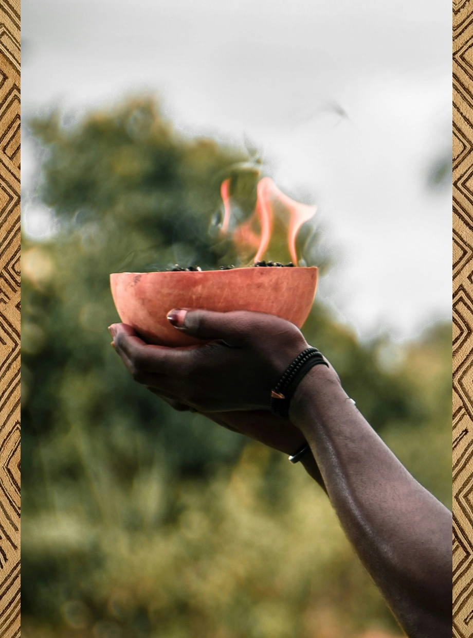 Two hands holding a wooden bowl with burning flames outdoors with blurred green background.