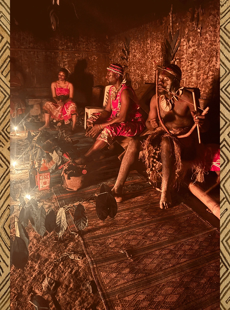 Three people in traditional attire sit indoors on woven mats, beside leaves and bottles near a fire.