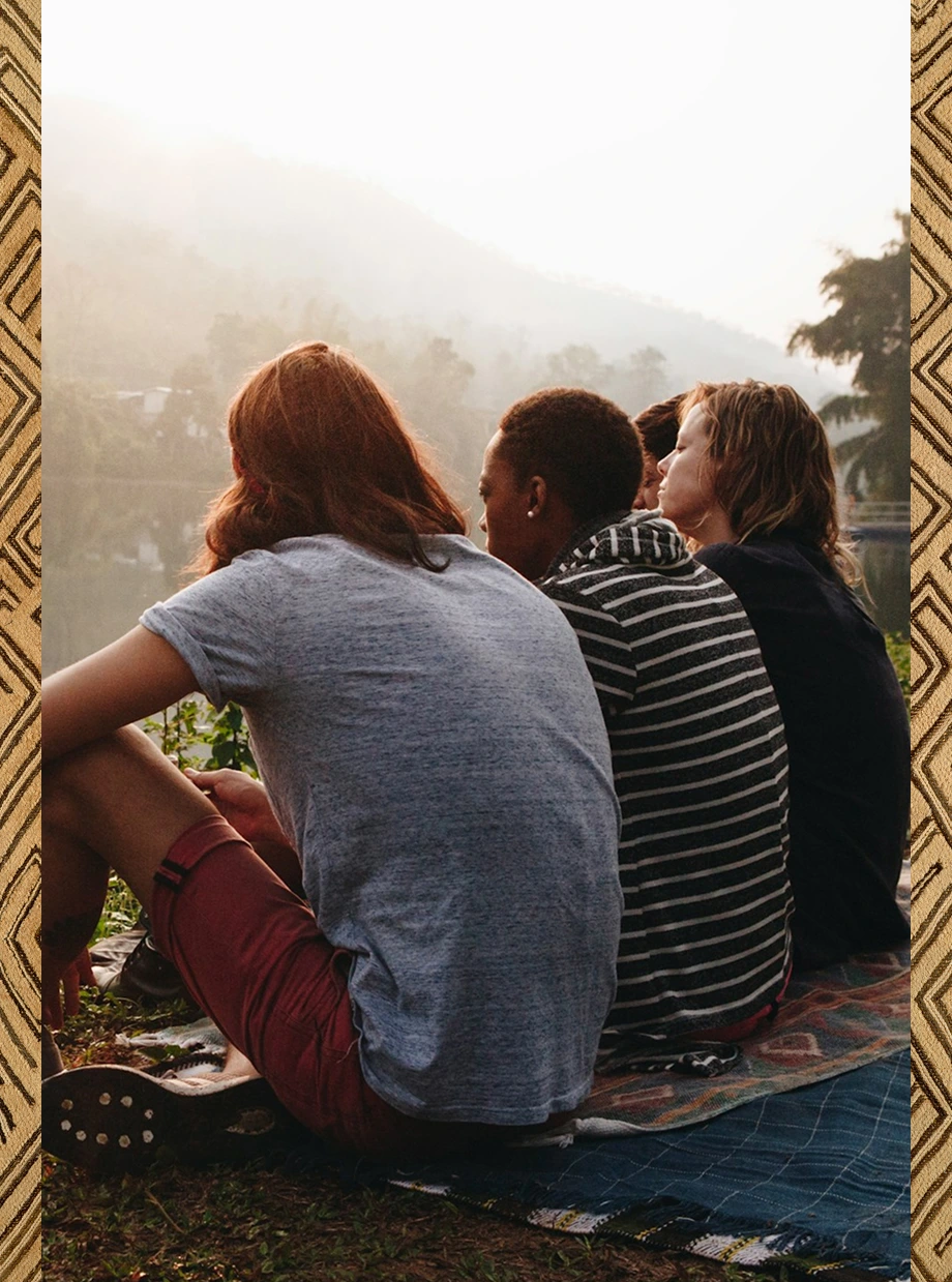 Four people sitting on blankets by a lake with mountains and trees in the background at sunset.
