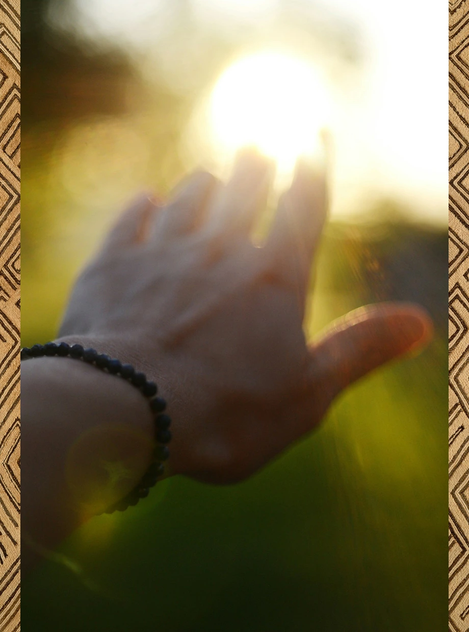 Hand with a beaded bracelet reaching toward bright sunlight in a blurred outdoor setting.