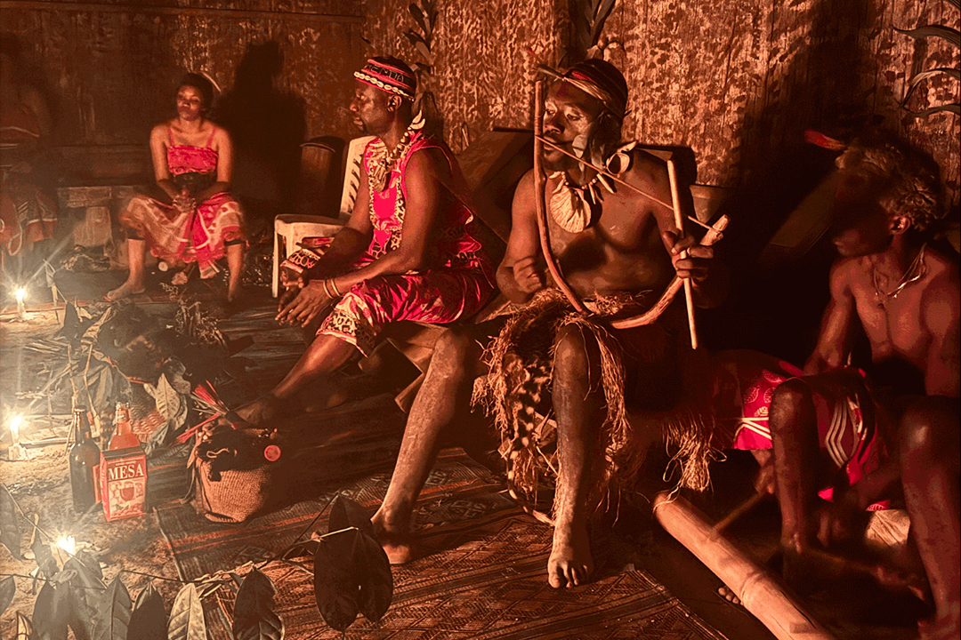 Four people in traditional attire sitting indoors near candlelight, one playing a musical instrument.