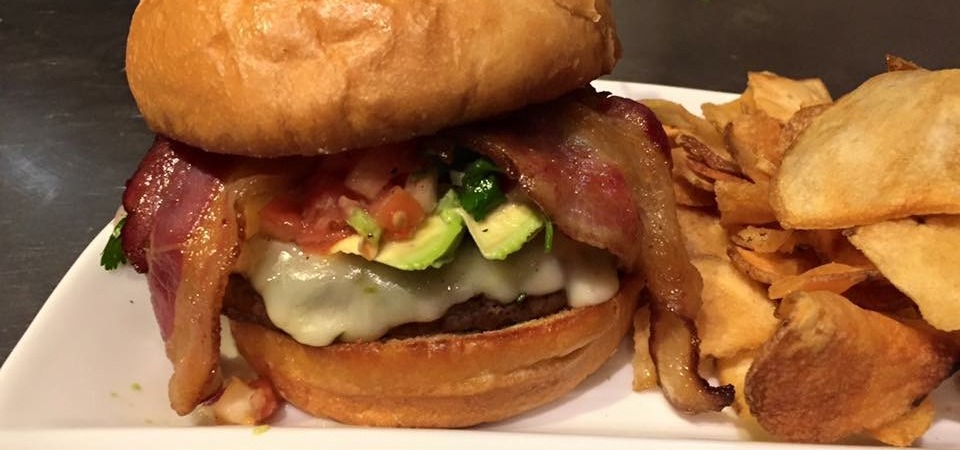 Bacon cheeseburger with avocado and tomato salsa served with thick potato chips on a white plate.