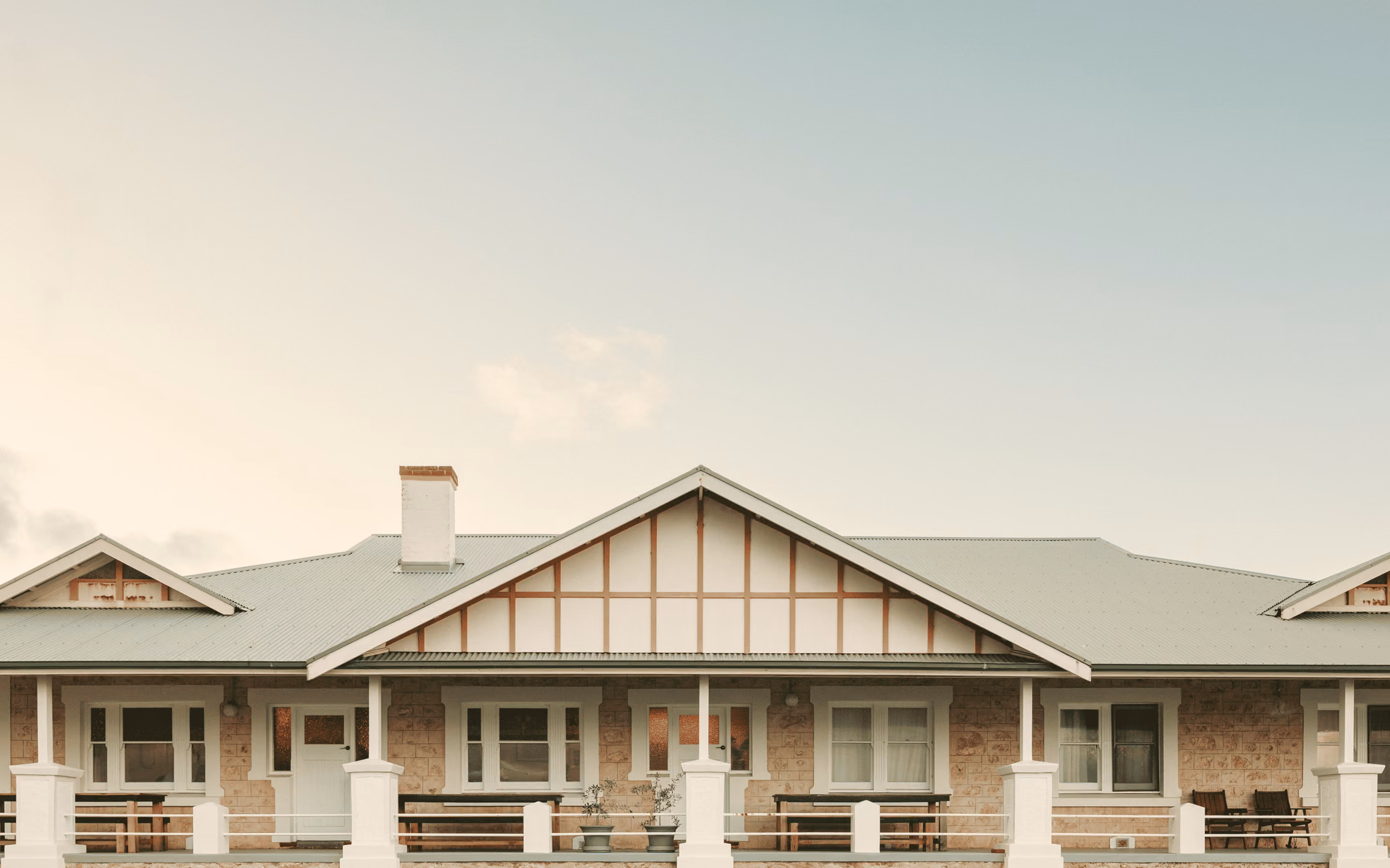 Stone house facade with symmetrical windows, white doors, porch benches, and a grey metal roof.
