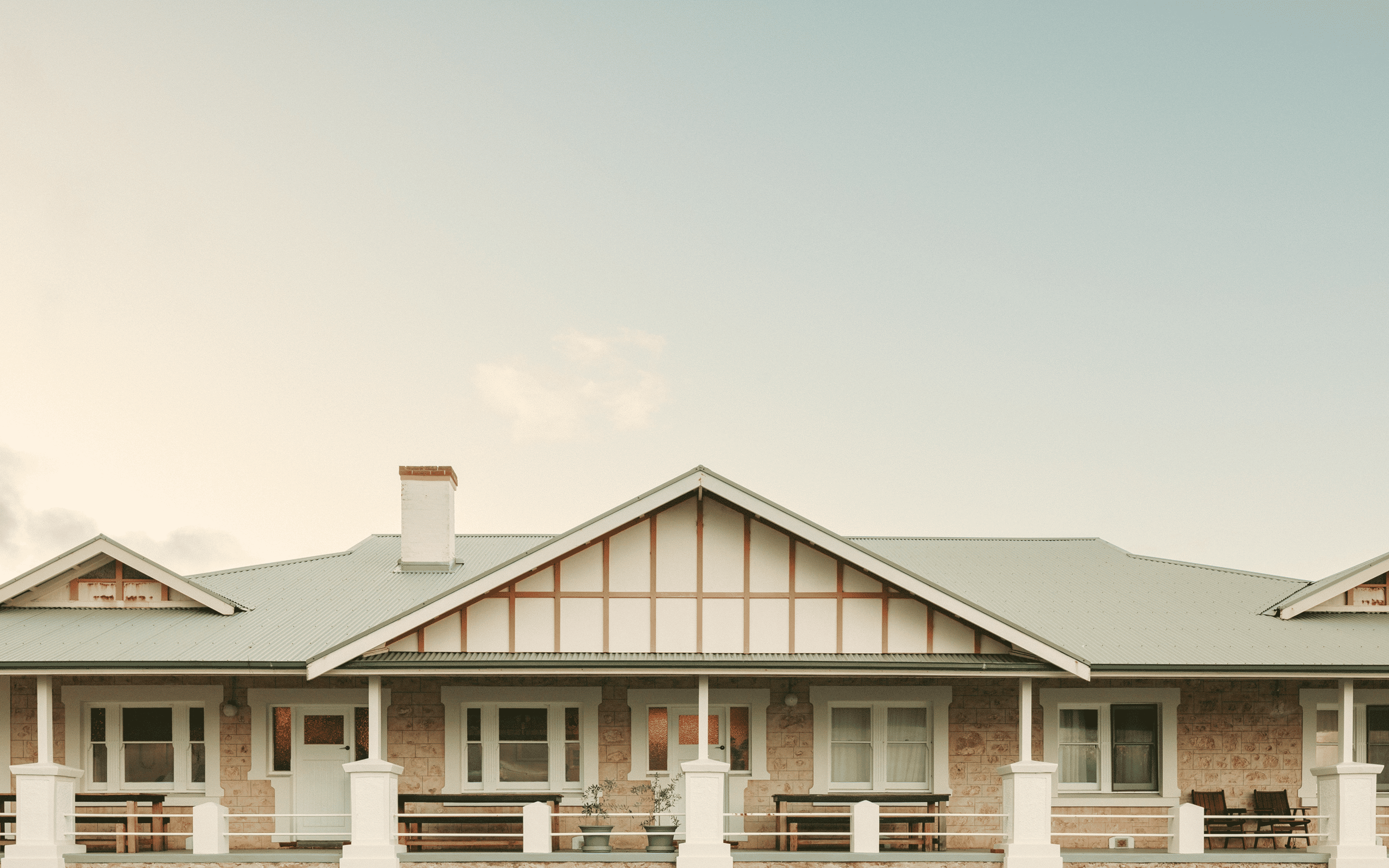 Stone house facade with symmetrical windows, white doors, porch benches, and a grey metal roof.