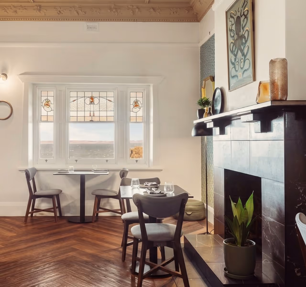 Bright dining area with wooden floor, two tables with chairs, stained glass window, and a dark tiled fireplace.