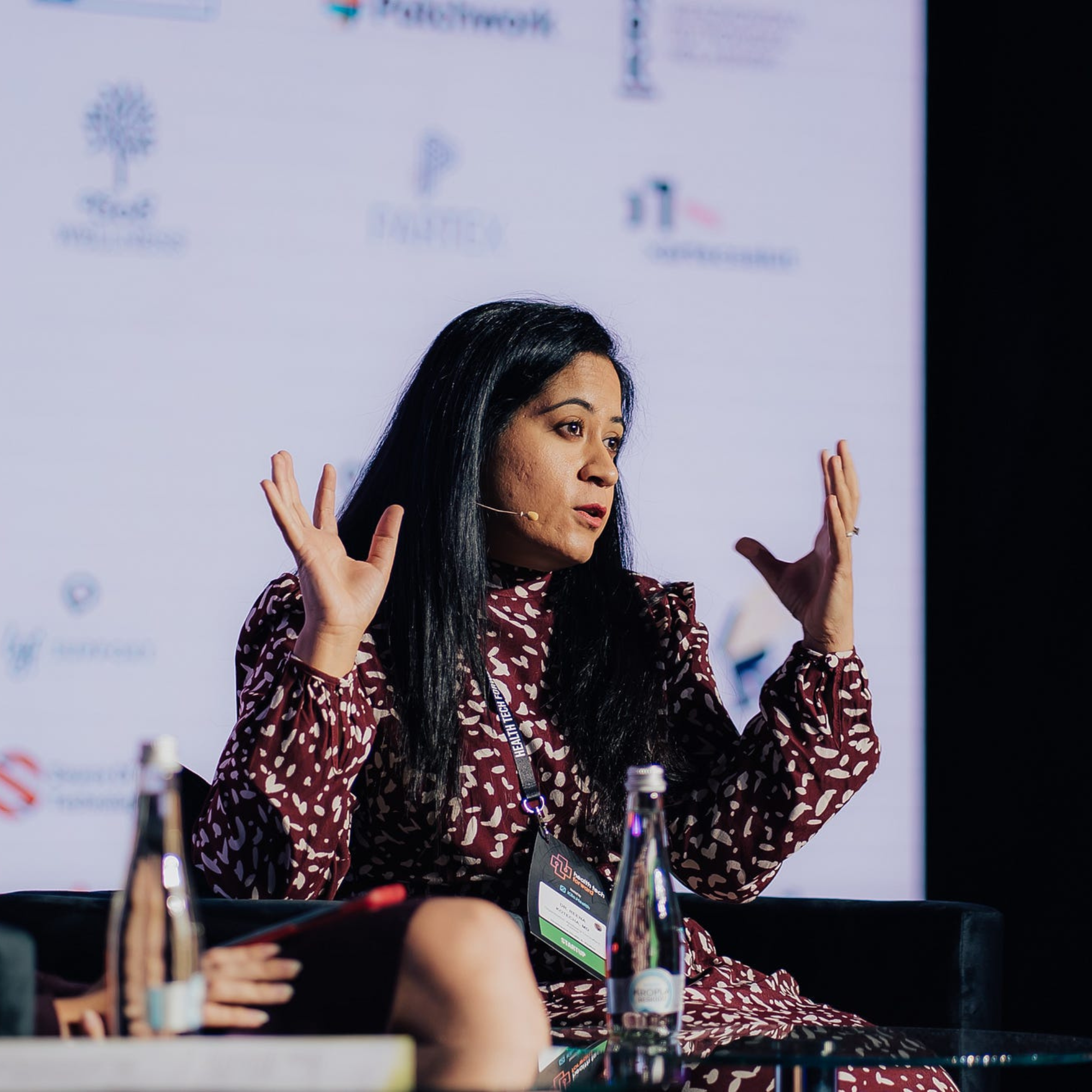 Woman with long black hair in maroon patterned dress speaking and gesturing with raised hands.