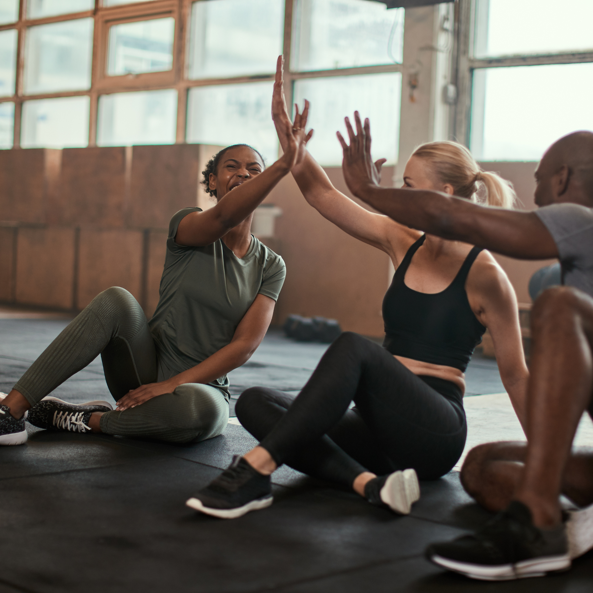 Three people sitting on gym floor giving high fives, smiling and celebrating workout success.