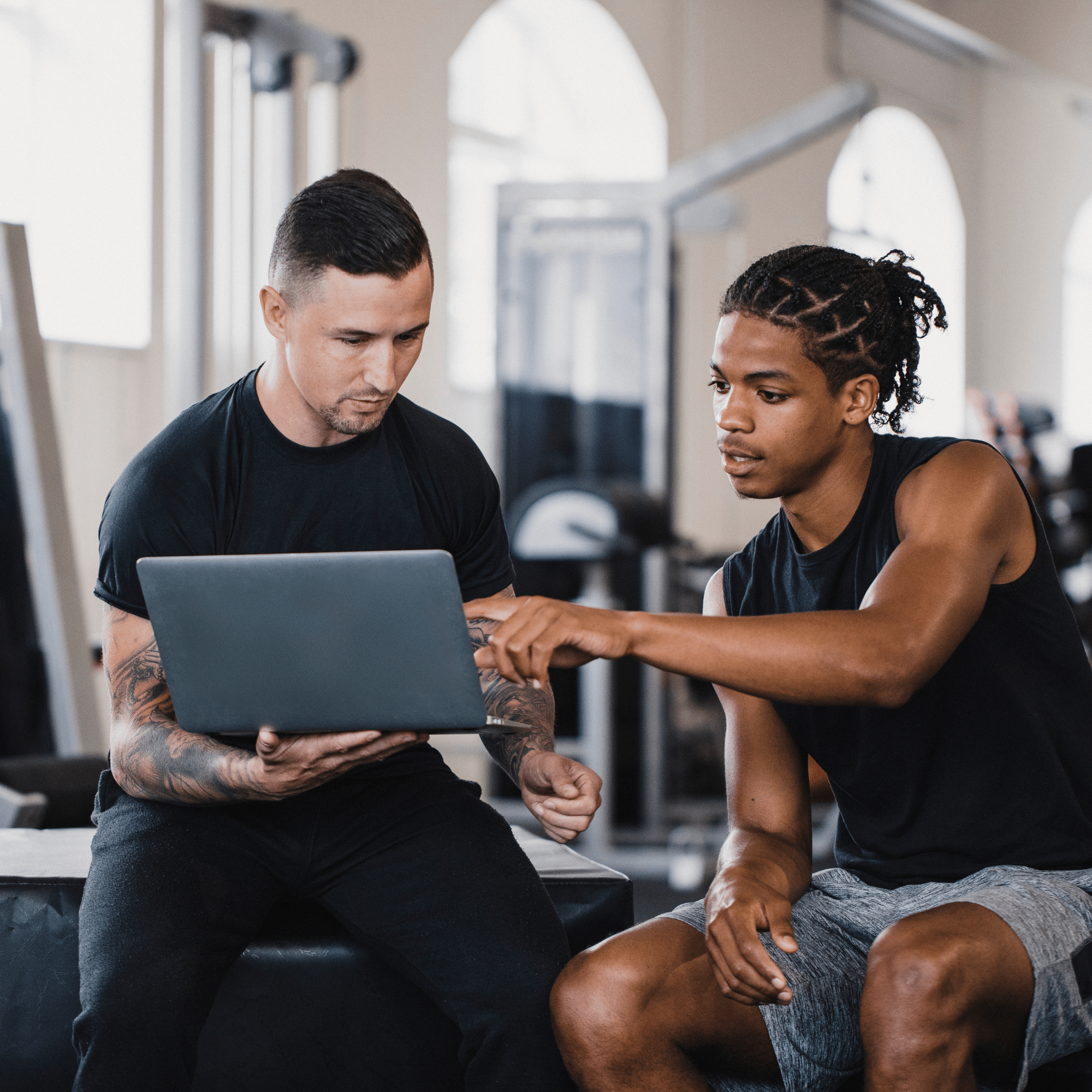 Two men in a gym looking at a laptop, one pointing at the screen while sitting on a bench.