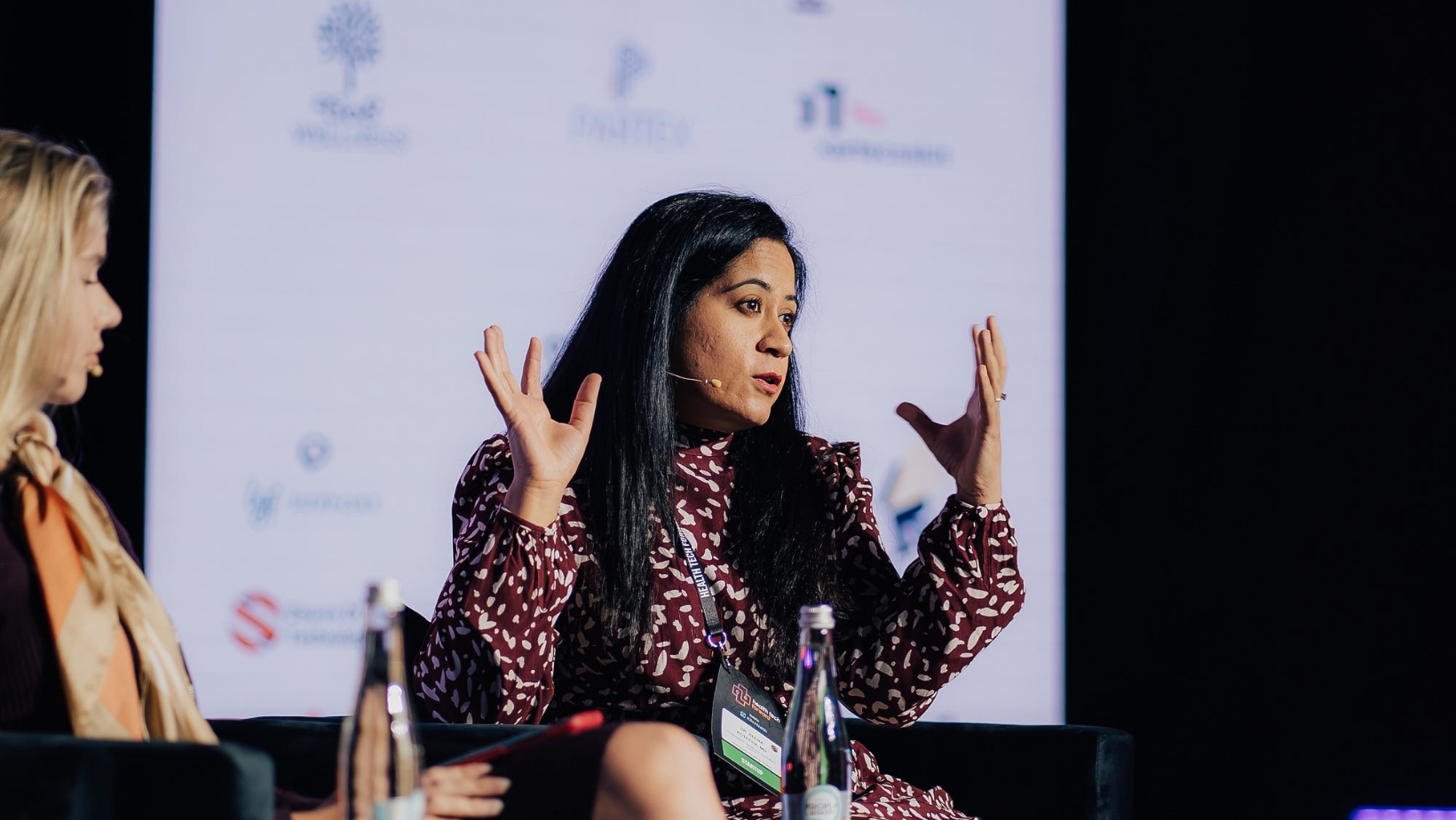 Woman speaking passionately on stage during a panel discussion with raised hands and microphone headset.