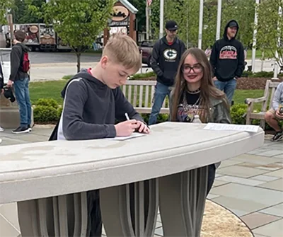 Boy writing on a paper at an outdoor table with a girl watching and people walking in background.