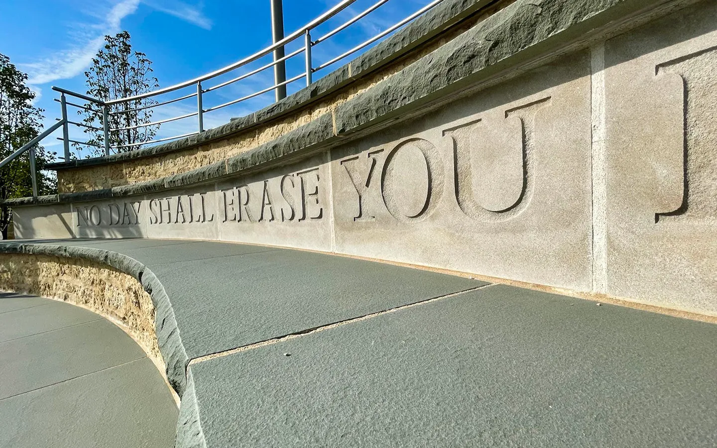 Curved stone memorial wall with carved text reading 'NO DAY SHALL ERASE YOU' and metal railings above.