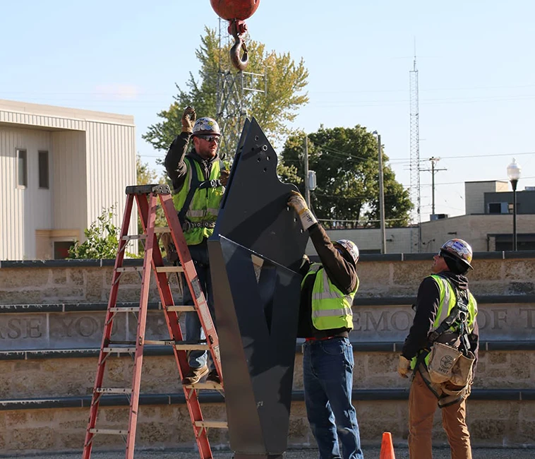 Construction workers in safety vests guiding a large black metal piece suspended by a crane hook.