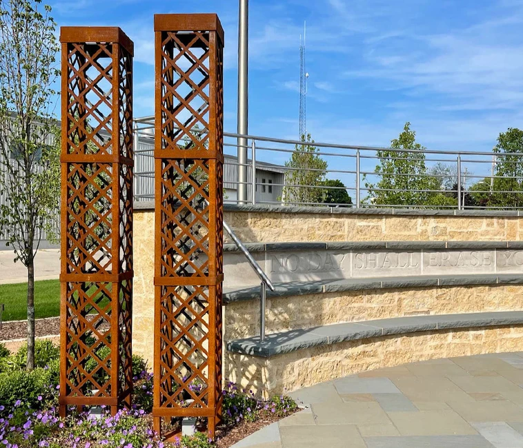 Two tall rust-colored metal lattice pillars near a curved stone wall with engraved text outdoors.