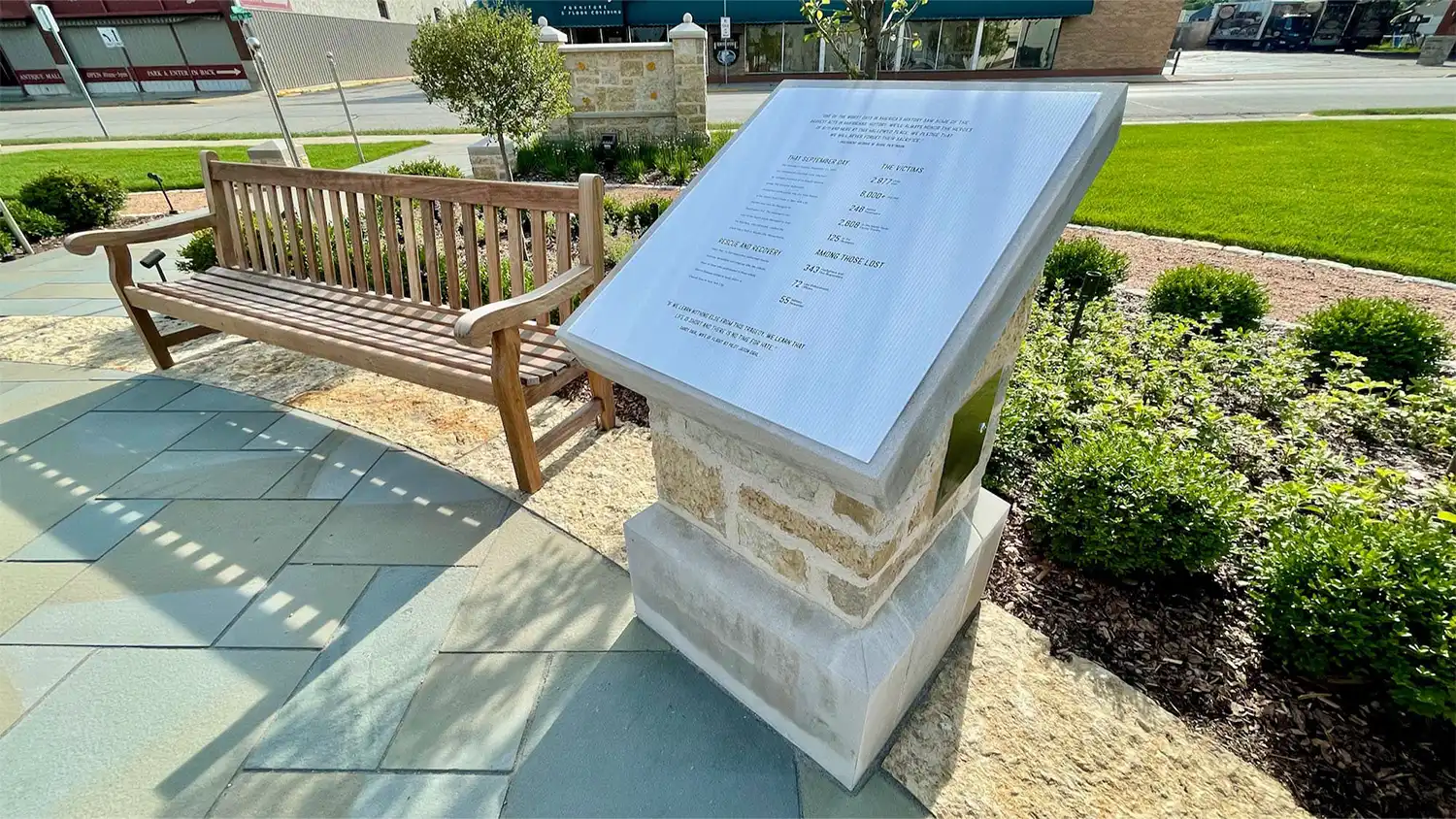 Stone plaque with engraved text near wooden bench in landscaped outdoor area with greenery.