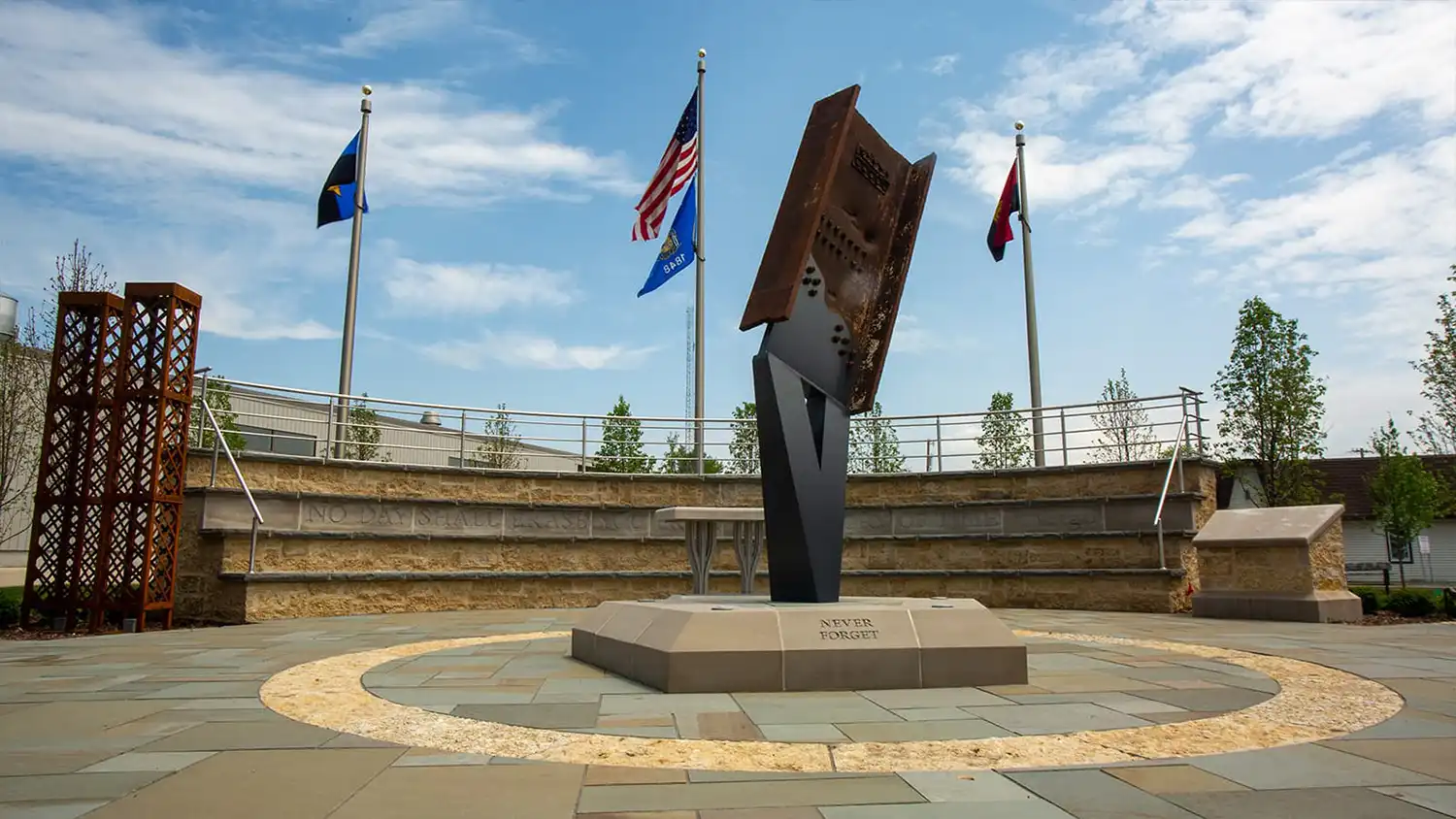 Outdoor memorial with rusted steel beam sculpture on a stone base inscribed 'Never Forget' with flags behind.