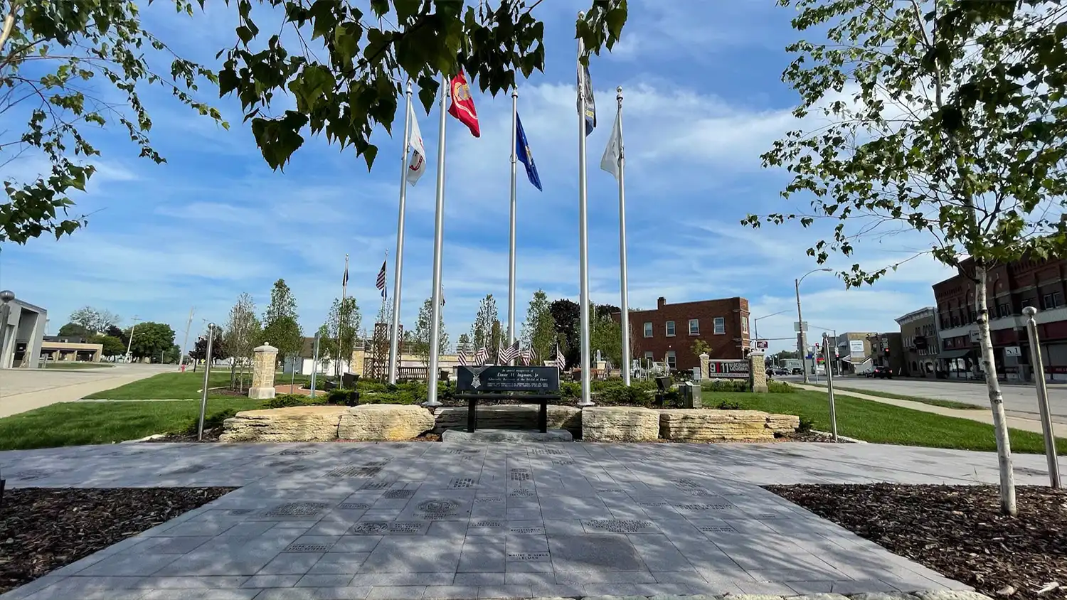 Veterans memorial with flagpoles, Memorial Day bench, and engraved stone walkway under blue sky.