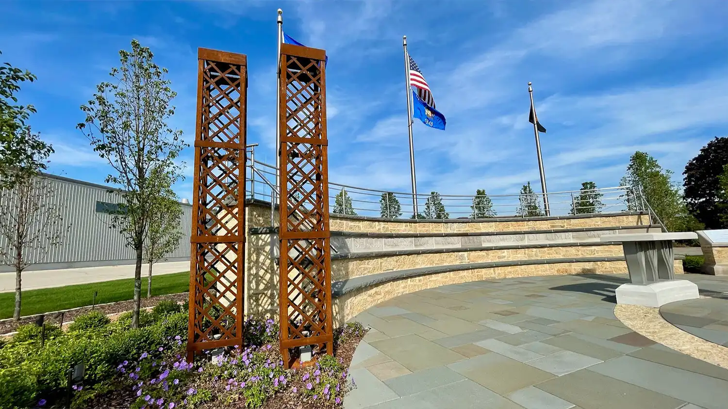 Rust-colored metal twin towers sculpture with flowers, flagpoles, and stone seating under blue sky.
