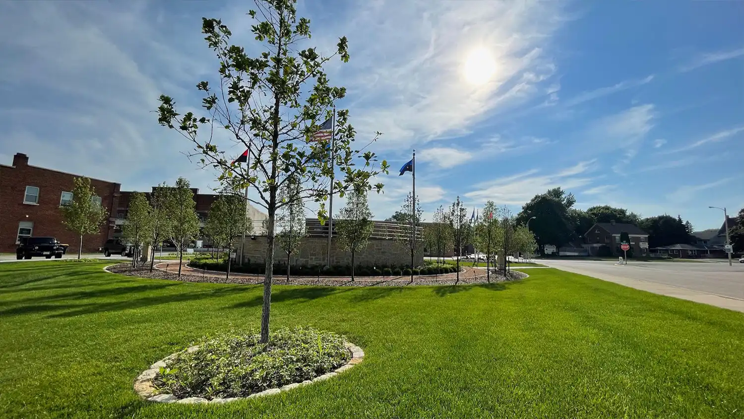 Sunny view of green lawn with young trees and flags near a brick building by an intersection.