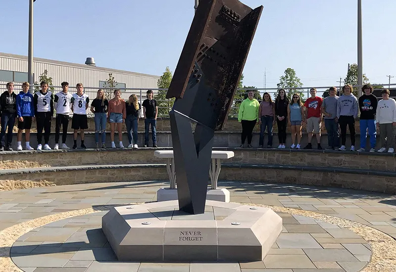 Memorial sculpture with 'Never Forget' inscription and people standing around a stone seating area.