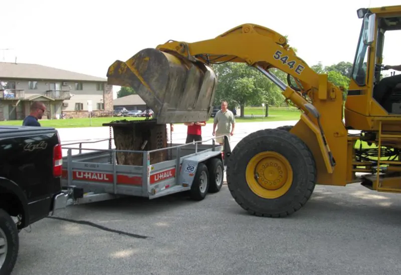 Yellow excavator lifting dirt bucket into U-Haul trailer attached to black truck with people nearby.