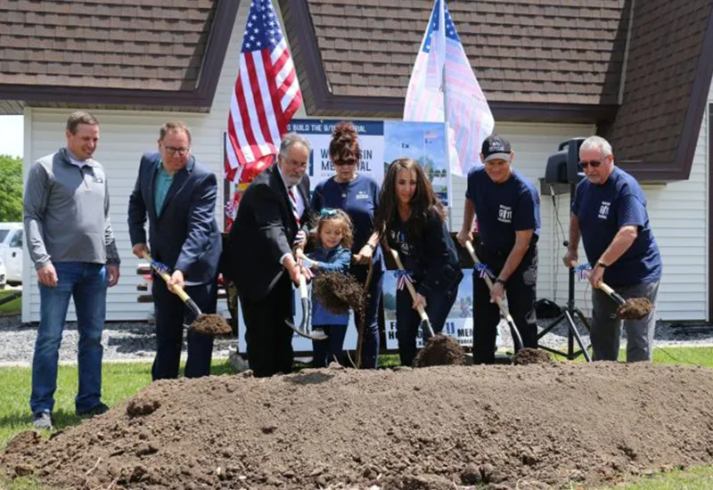 Group of people at groundbreaking ceremony holding shovels with American flags in background.