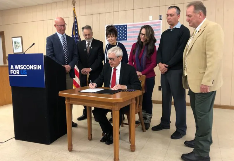 Man in suit signs document at table with six people standing behind and an American flag nearby.