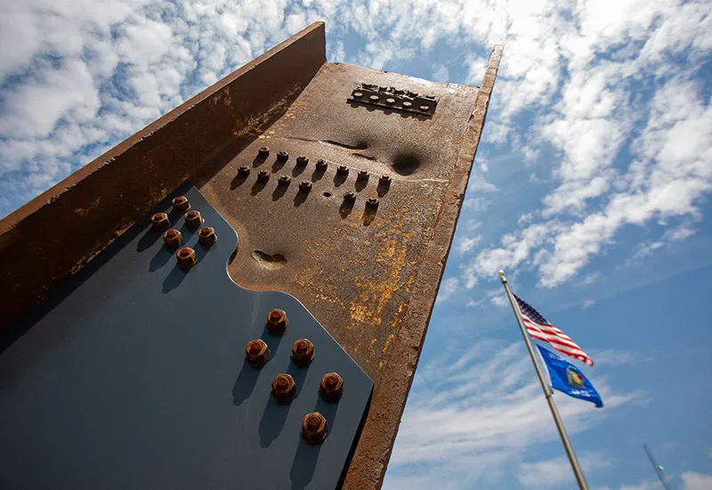 Rusty steel beam with bolts against a partly cloudy sky and an American flag in the background.