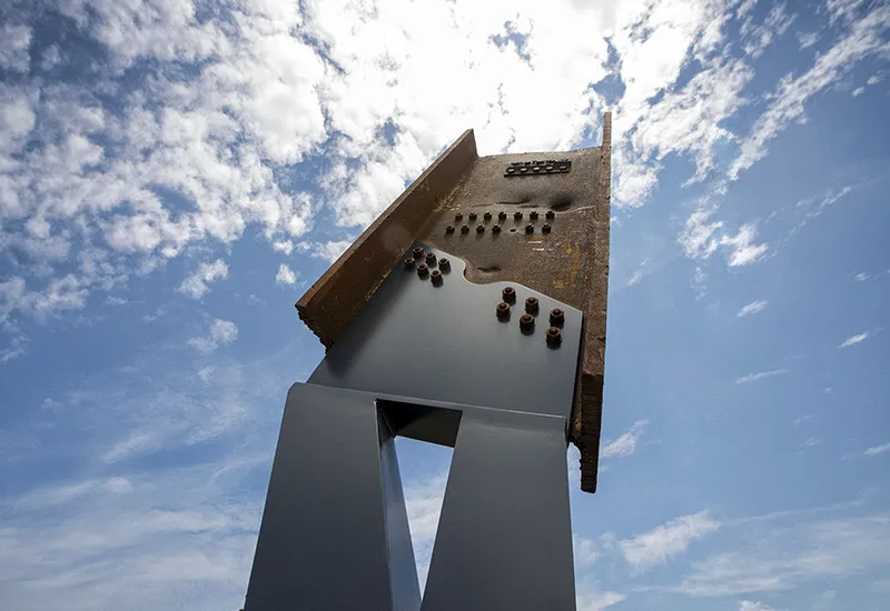 Close-up angled view of a large metal sculpture bolted and weathered, against a blue sky with scattered clouds.
