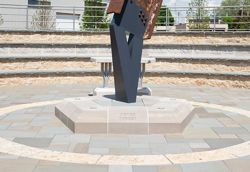 Steel and stone memorial with 'Never Forget' inscription on stone base outdoors.