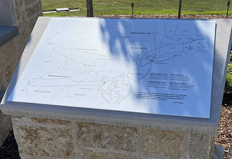 Outdoor stone plaque map with flight routes and text, partially in shadow, on a grassy lawn background.