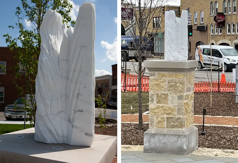 White marble sculpture with vertical, textured surfaces displayed on a stone pedestal outdoors in town.