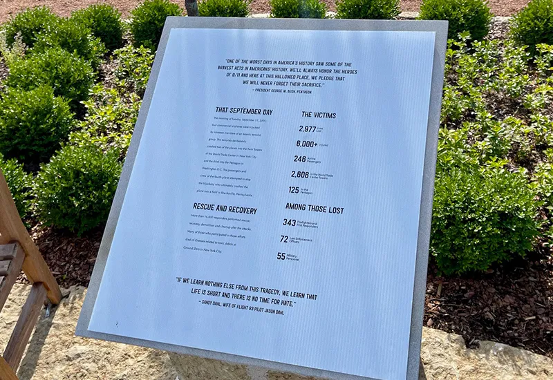 Memorial plaque with September 11 victims stats and quotes surrounded by shrubs and stone.