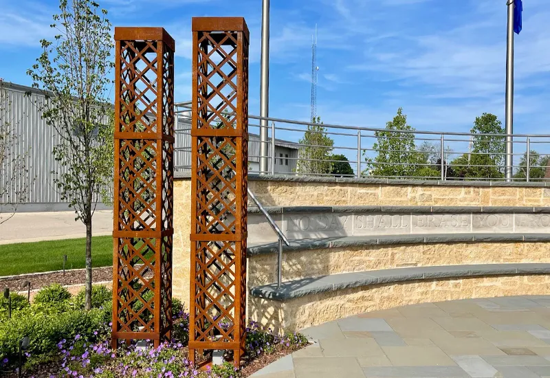 Two rusty metal lattice trellises stand in a garden bed near a curved stone wall with engraved text outdoors.