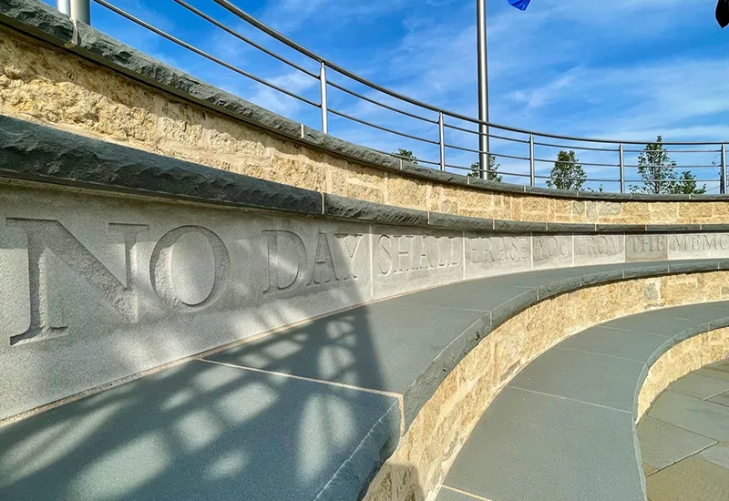 Curved stone bench wall engraved with 'NO DAY SHALL ERASE YOU FROM THE MEMO' under blue sky and flag.