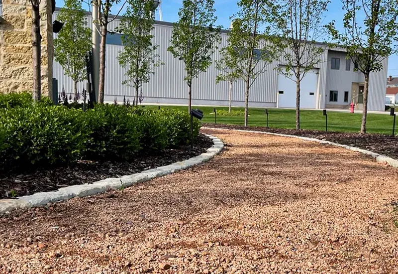 Gravel pathway bordered by stone edging, bushes, and trees leading to a light industrial building.
