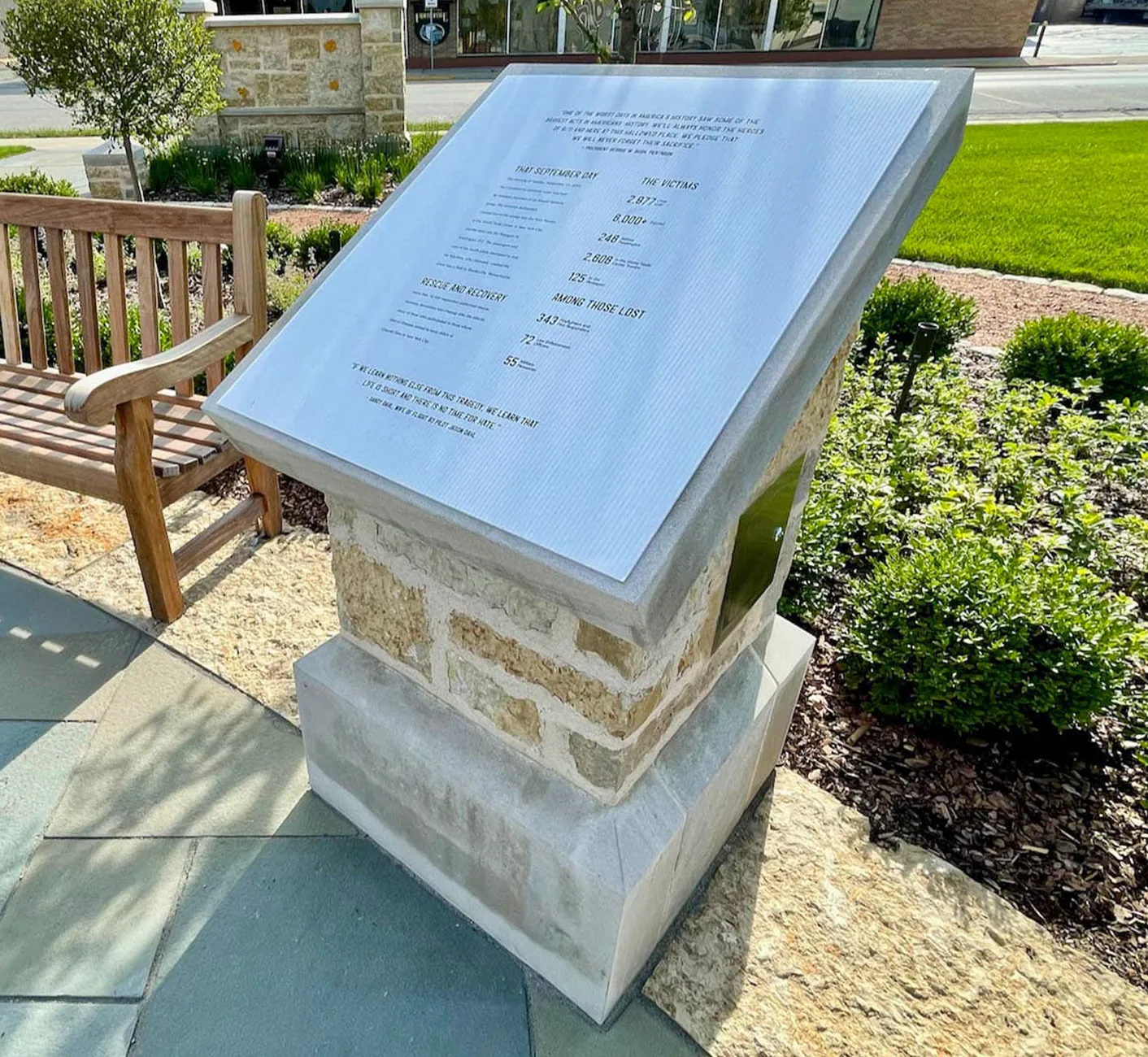 Stone memorial plaque with details about victims and rescue on September 11, placed outdoors by a wooden bench.