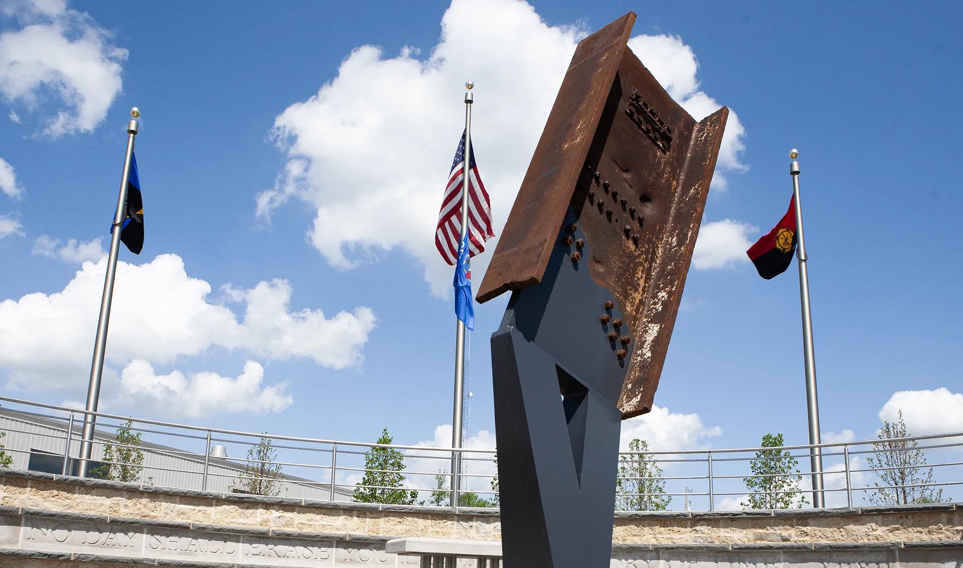 Steel beam sculpture with bolts in outdoor memorial setting under blue sky with clouds.