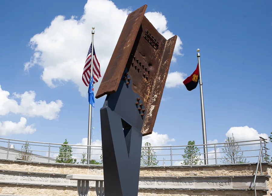 Steel beam sculpture with bolts in outdoor memorial setting under blue sky with clouds.
