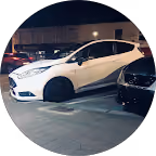 White car with black wheels parked at night between two other cars in a lot.