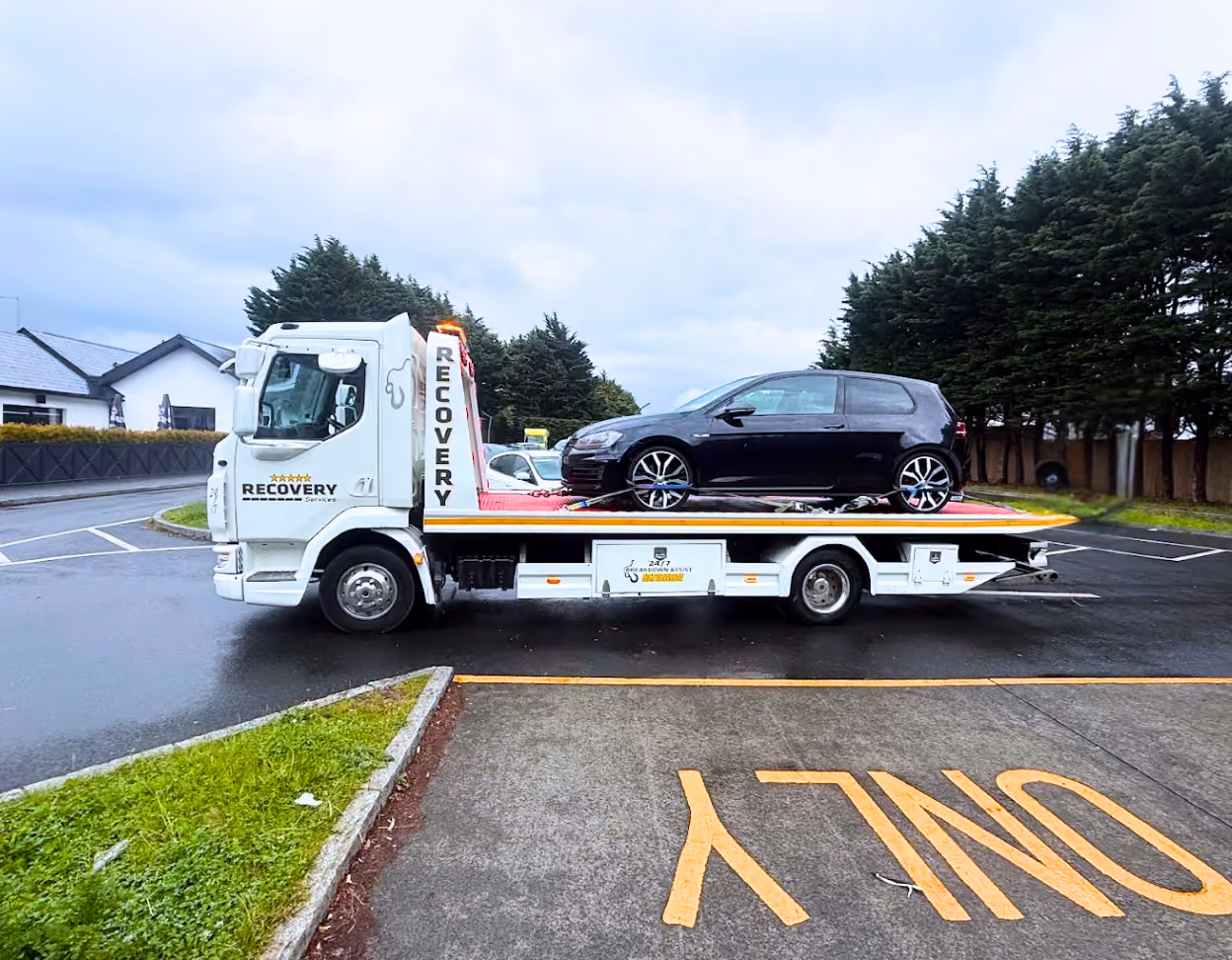 White recovery truck carrying a black hatchback car in a parking lot with grass and trees.