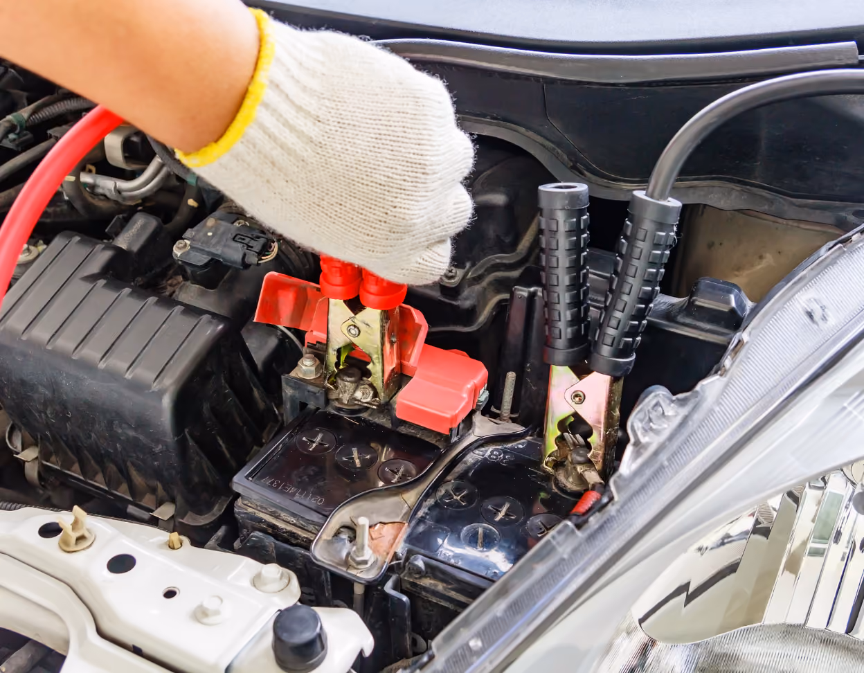 Gloved hand attaching red jumper cable to car battery terminal under open hood.