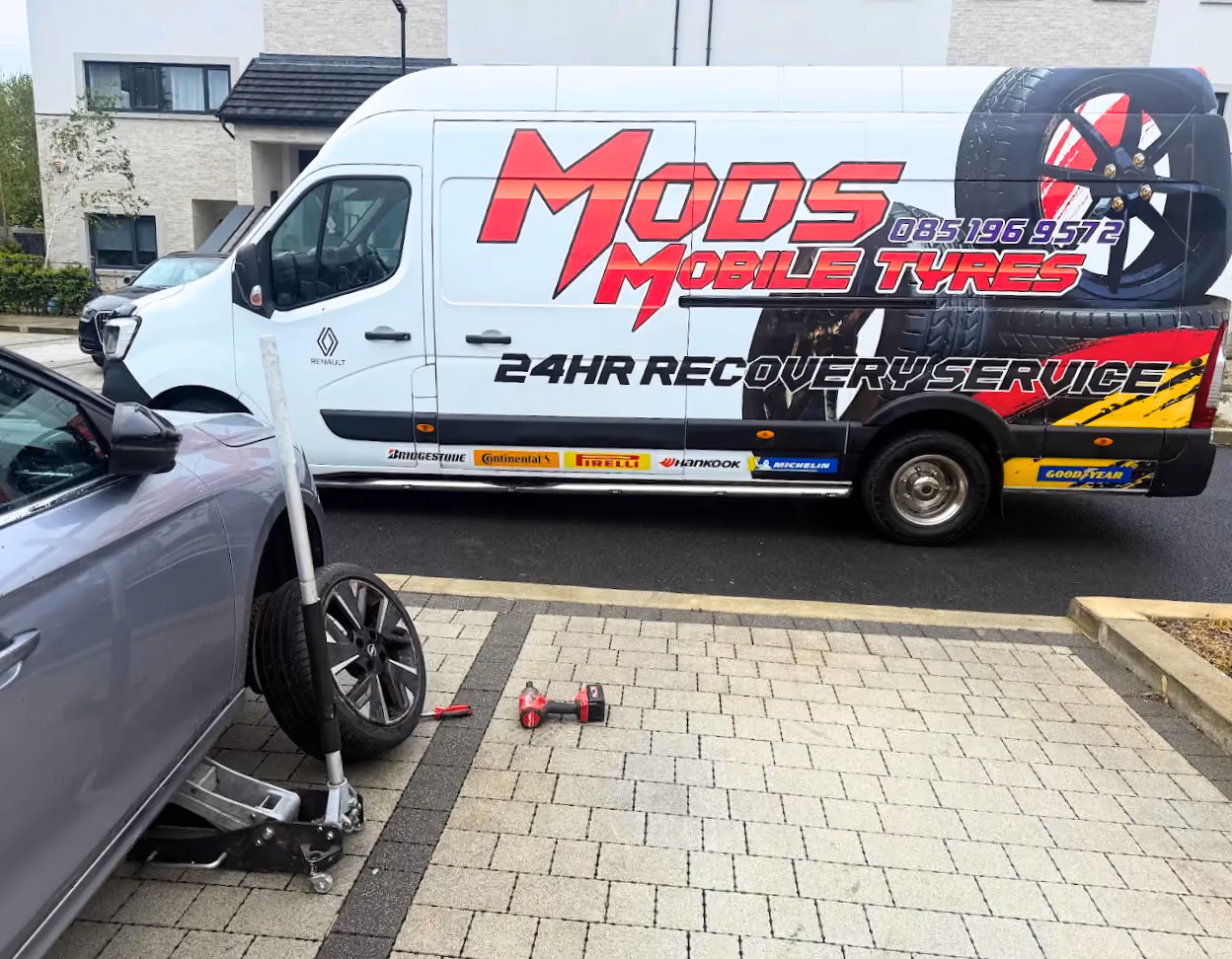White van with 'Mods Mobile Tyres 24HR Recovery Service' text parked beside a car with a tire change in progress.
