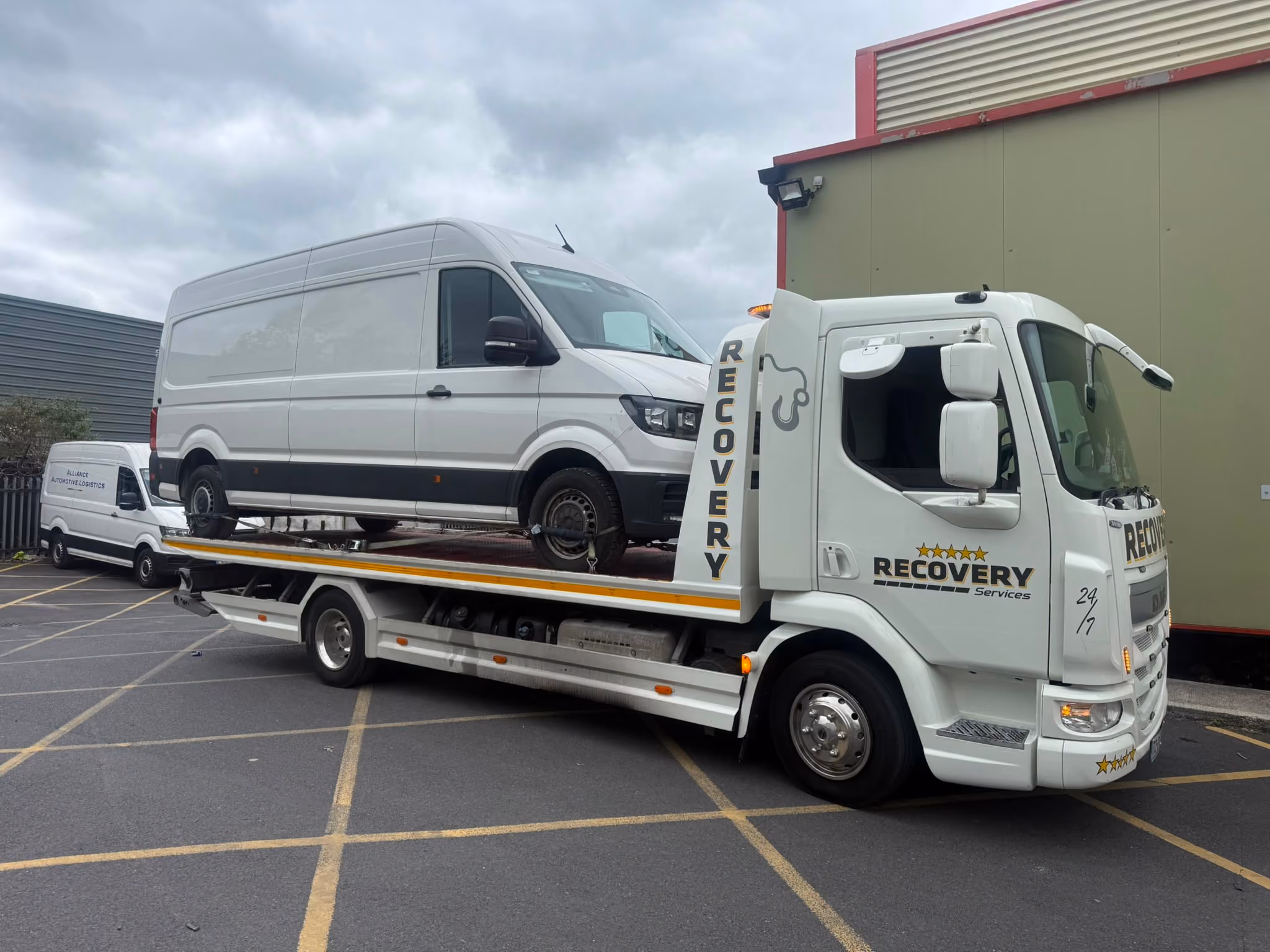 Tow truck with Recovery Services logo carrying a white van in a parking lot on a cloudy day.