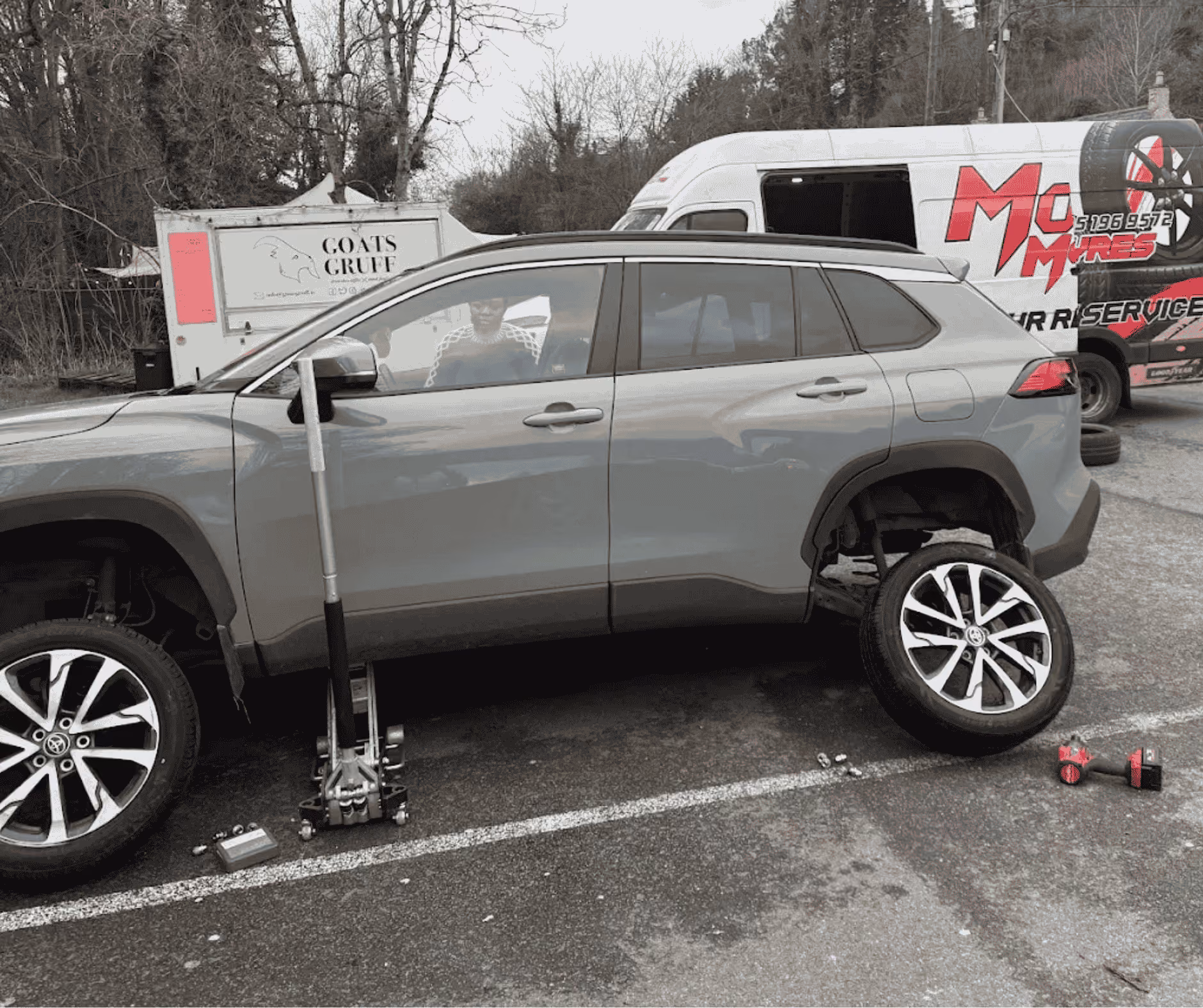Gray SUV with rear right wheel removed, jack and tools beside it, and service van in background.