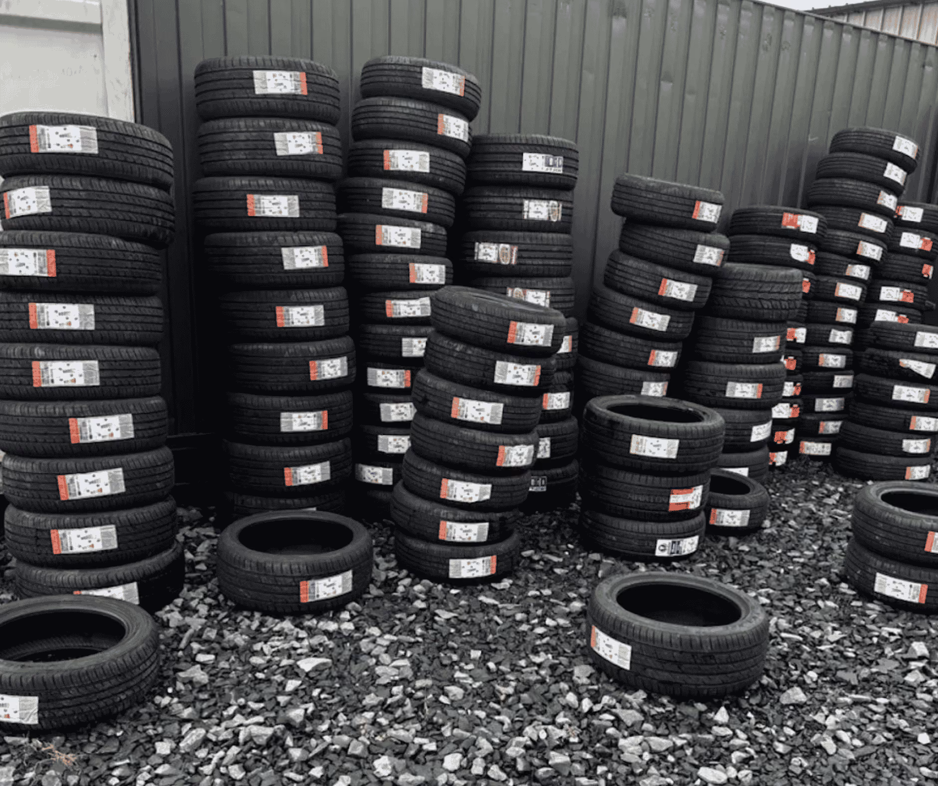 Stacks of new black tires with white labels arranged outdoors on rocky ground near a fence.