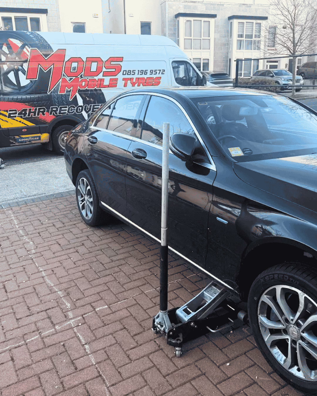 Black car raised on a jack with a Mods Mobile Tyres van parked behind it.