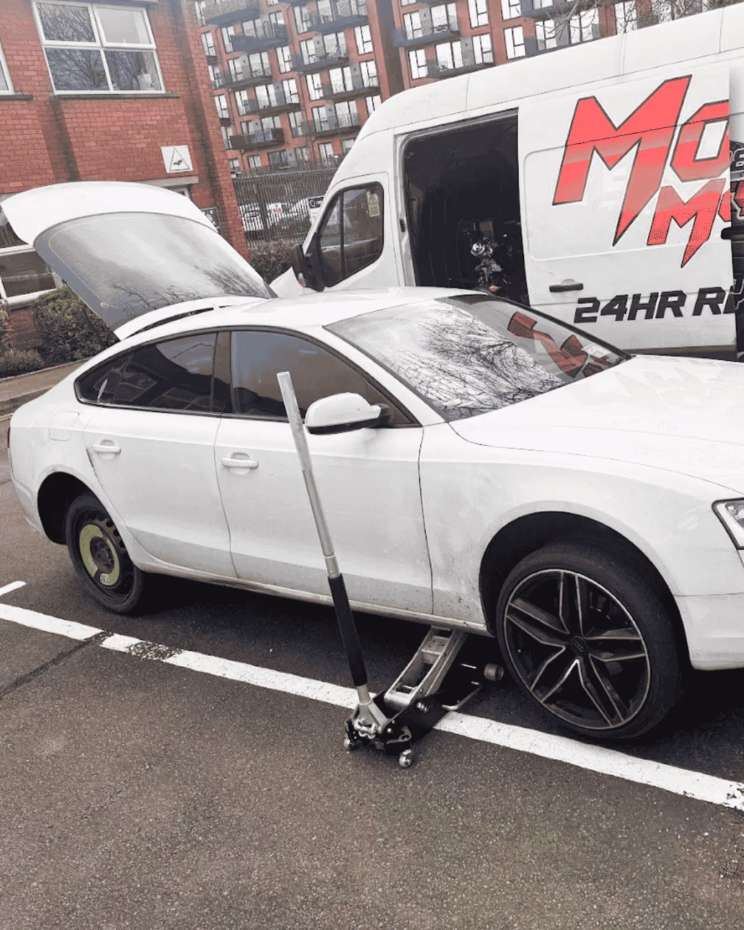 White car with a flat rear tire lifted by a jack in a parking lot next to a white van.