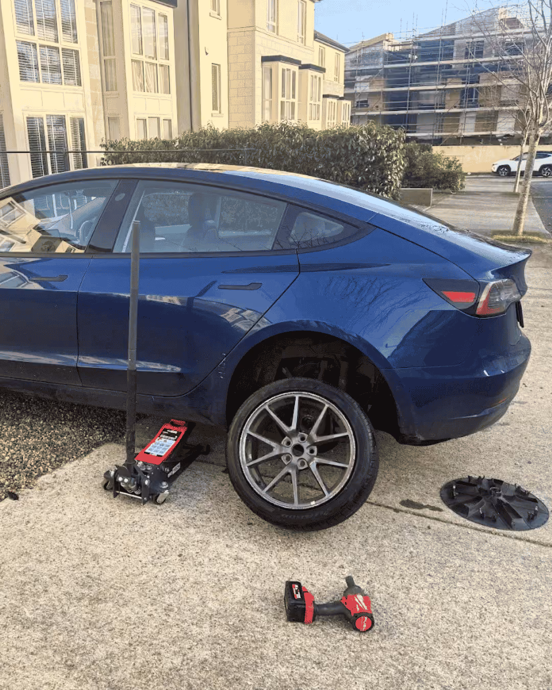 Blue car raised on jack with rear wheel removed, tire and hubcap on ground nearby and a handheld tool.