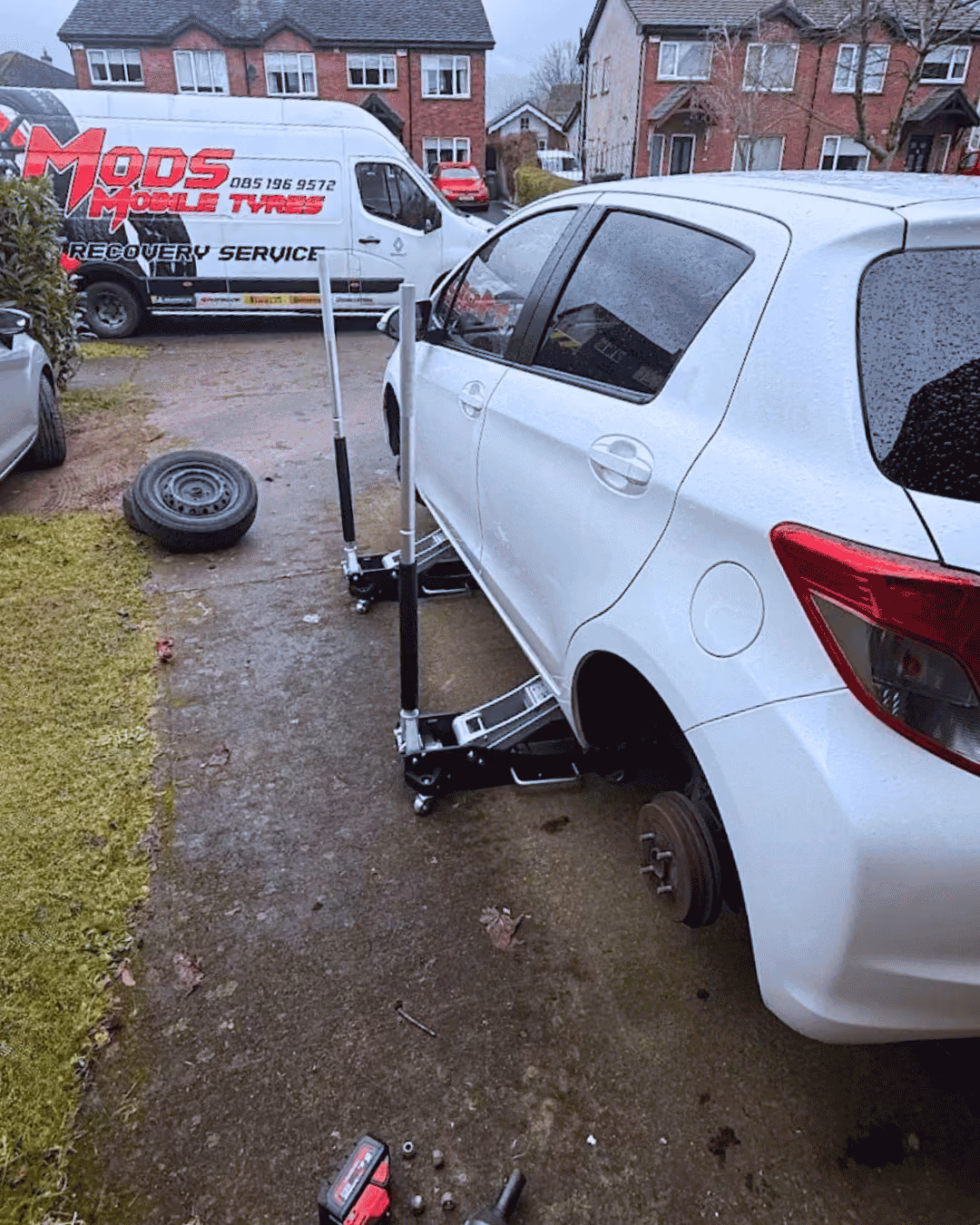 White car on jacks with a removed rear wheel and a tire laying nearby on pavement.